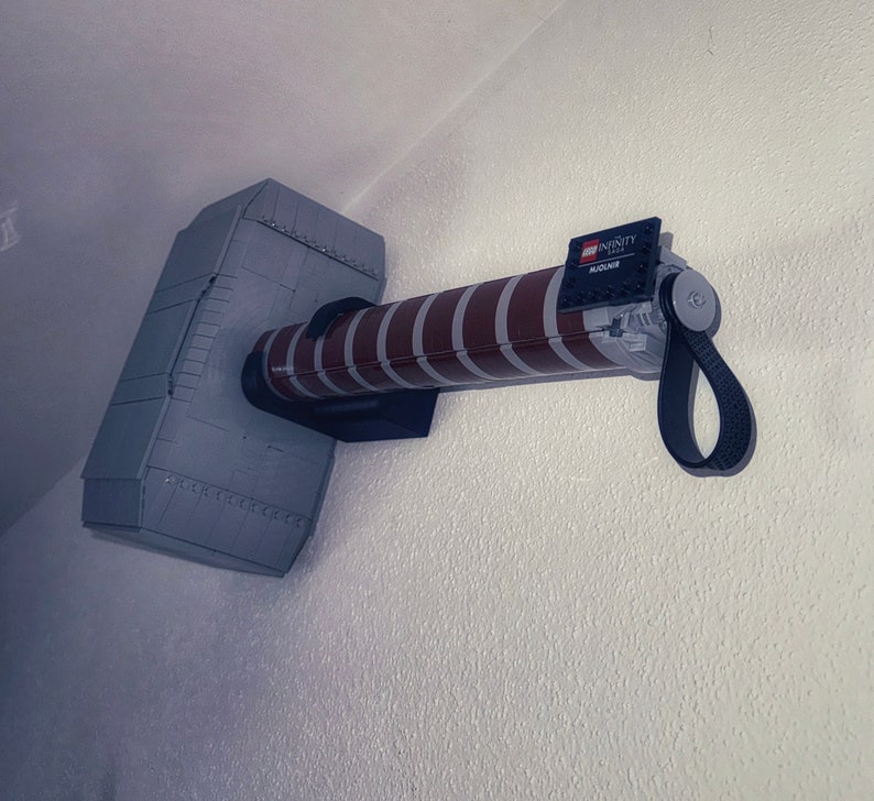 Op de afbeelding: Een grijze en bruine LEGO Mjolnir-hamer, gemonteerd aan een witte muur. De hamerkop is grijs met klinknageldetails en de steel is bruin met witte strepen. De steel heeft een zwarte band en het LEGO Infinity-logo.