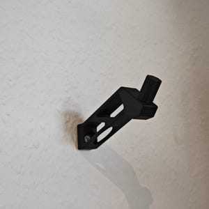 May include: A black wall bracket with a geometric design, attached to a white wall with a screw. The bracket has a rectangular base with two angled cutouts and a cylindrical post extending from the top.