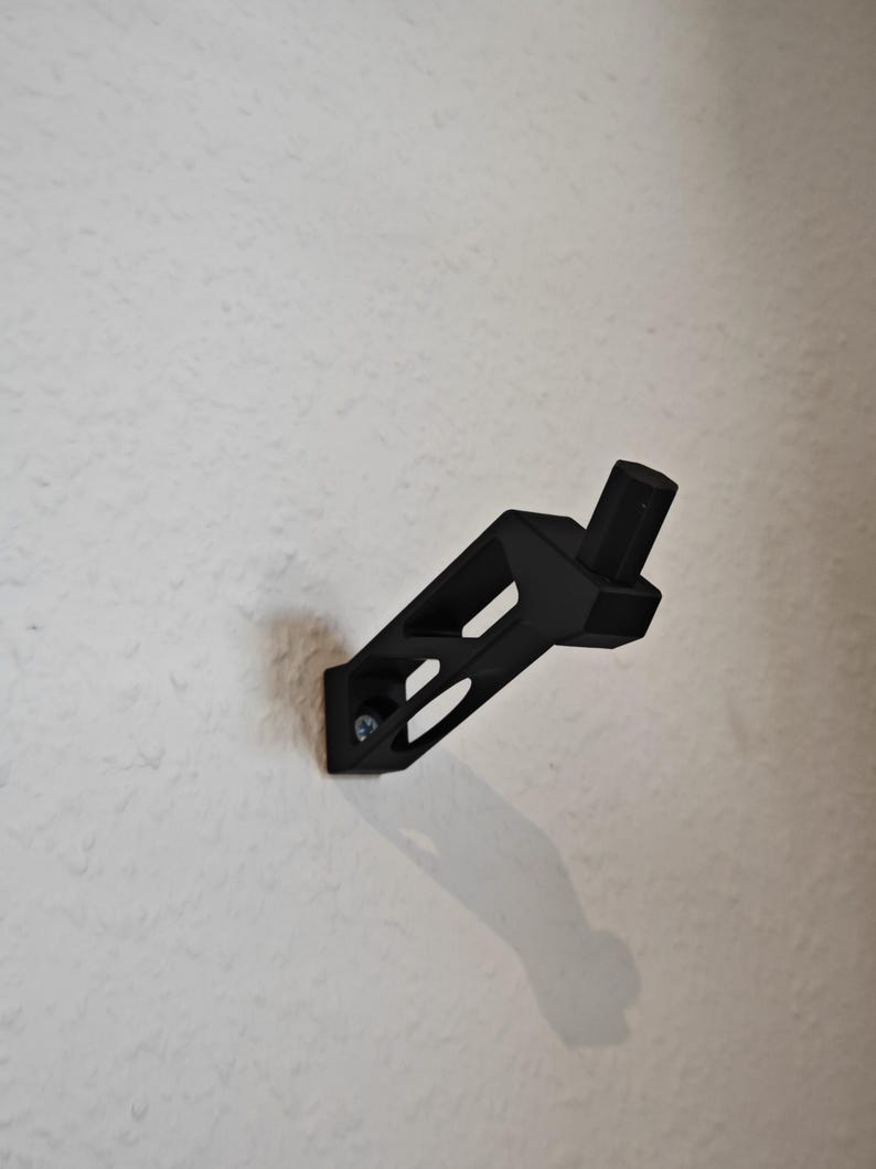 May include: A black wall bracket with a geometric design, attached to a white wall with a screw. The bracket has a rectangular base with two angled cutouts and a cylindrical post extending from the top.