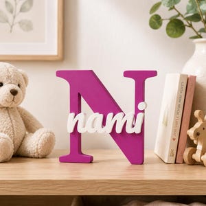 May include: A freestanding decorative letter N in a vibrant magenta color, featuring the name nami in white lowercase script lettering layered across the center of the letter. The object is constructed with a smooth, solid finish and stands upright.