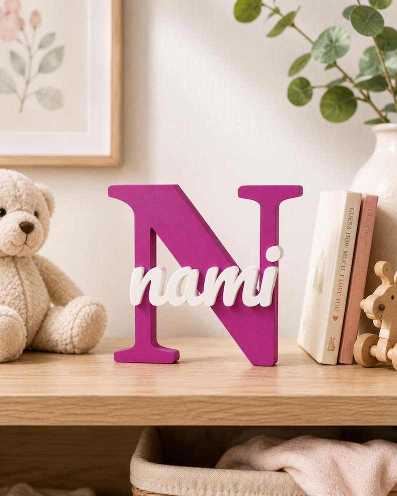 May include: A freestanding decorative letter N in a vibrant magenta color, featuring the name nami in white lowercase script lettering layered across the center of the letter. The object is constructed with a smooth, solid finish and stands upright.