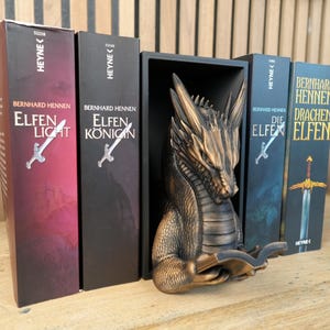 May include: A bronze dragon bookend holds several fantasy novels with titles like "Elfen Light" and "Die Elfen". The books have sword illustrations and are in a row on a wooden surface. A small potted plant is in the background.
