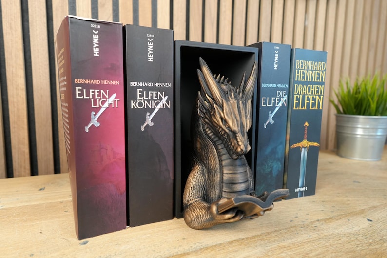 May include: A bronze dragon bookend holds several fantasy novels with titles like "Elfen Light" and "Die Elfen". The books have sword illustrations and are in a row on a wooden surface. A small potted plant is in the background.