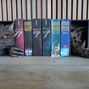 May include: A dragon-themed bookshelf with a gray dragon head and tail, holding several books. The books have colorful covers with titles in German. The dragon has red eyes and sharp teeth.