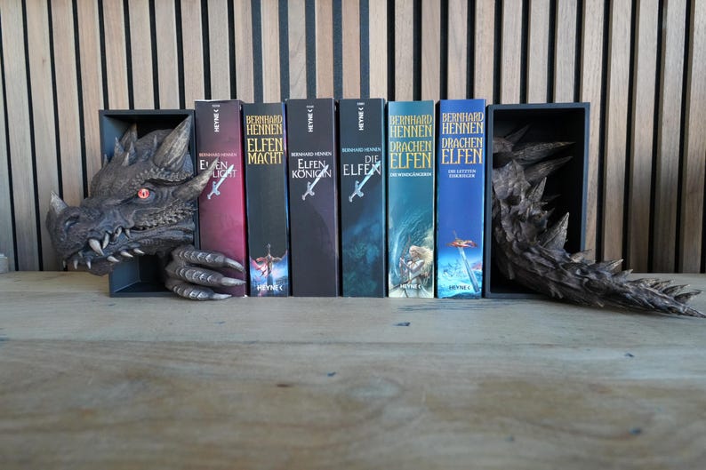 May include: A dragon-themed bookshelf with a gray dragon head and tail, holding several books. The books have colorful covers with titles in German. The dragon has red eyes and sharp teeth.
