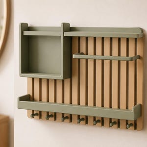 May include: A wall-mounted organizer featuring a light brown slatted back panel with olive green accents. It includes a top shelf, a deep storage box, a horizontal bar, a bottom ledge, and eight small pegs along the base for hanging items.