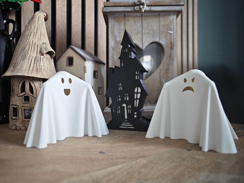 Puede incluir: Dos figuritas de fantasmas blancas con diferentes expresiones faciales. El fantasma de la izquierda sonr&iacute;e y el fantasma de la derecha frunce el ce&ntilde;o. Los fantasmas est&aacute;n sentados sobre una superficie de madera frente a una silueta negra de una casa encantada.