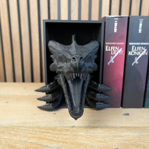 May include: A black dragon head sculpture with an open mouth, displayed in a black box. The sculpture is next to several books with the title "Elfen Licht" and "Elfen Konigin". The background is a wooden wall.