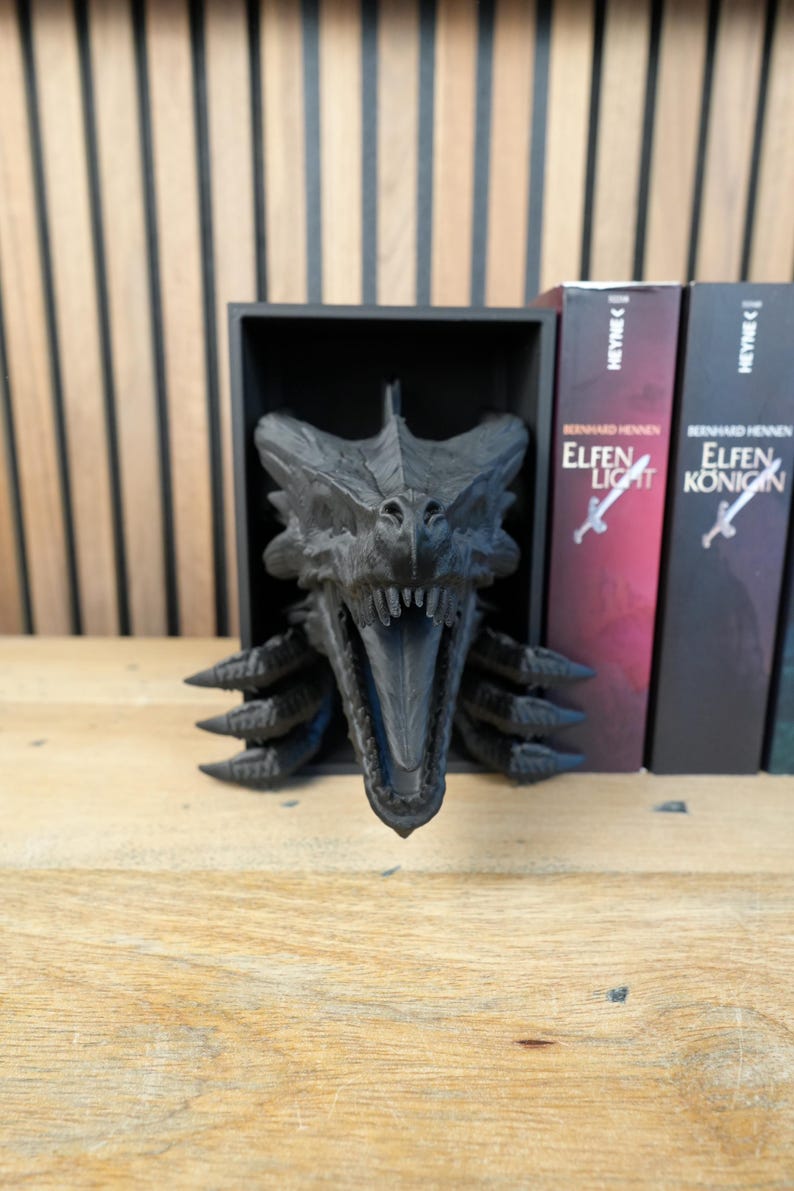 May include: A black dragon head sculpture with an open mouth, displayed in a black box. The sculpture is next to several books with the title "Elfen Licht" and "Elfen Konigin". The background is a wooden wall.