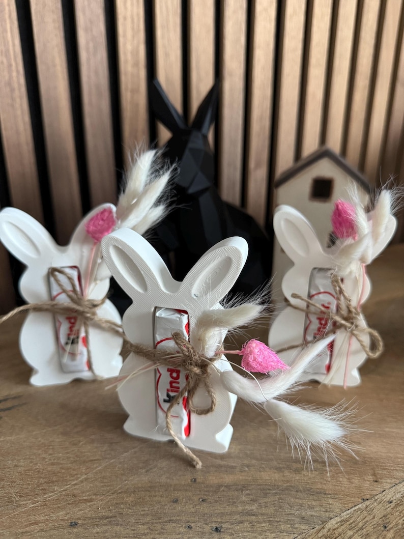 May include: Three white ceramic bunny figurines with a chocolate bar inside each. The bunnies are decorated with brown twine and white and pink dried flowers.