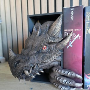 May include: A detailed dragon head bookend in shades of gray and bronze. The dragon's head, with sharp teeth and a menacing gaze, emerges from a black rectangular base. The bookend is positioned between two books, adding a fantasy element to a bookshelf.