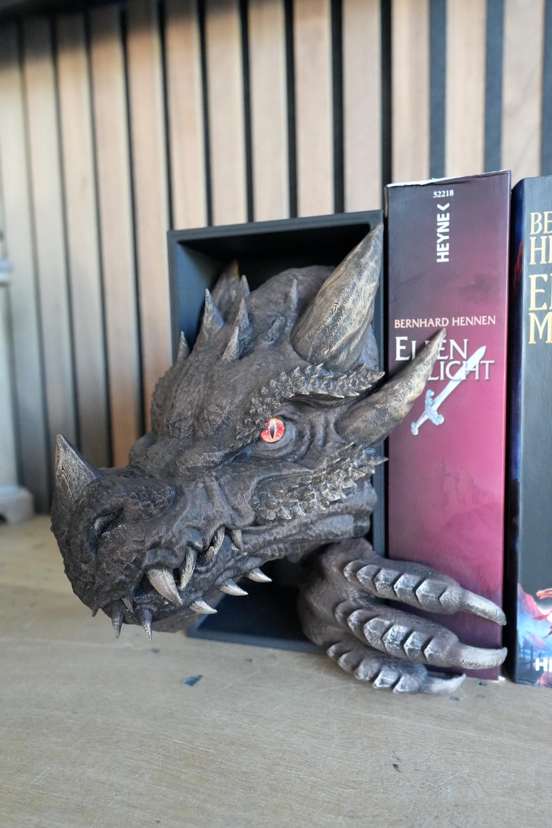 May include: A detailed dragon head bookend in shades of gray and bronze. The dragon's head, with sharp teeth and a menacing gaze, emerges from a black rectangular base. The bookend is positioned between two books, adding a fantasy element to a bookshelf.