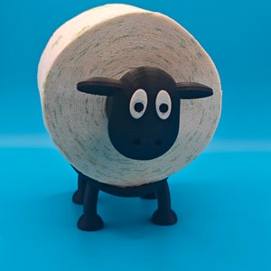 May include: A black sheep-shaped toilet paper holder with a white roll of toilet paper. The sheep has a black face with two white eyes.