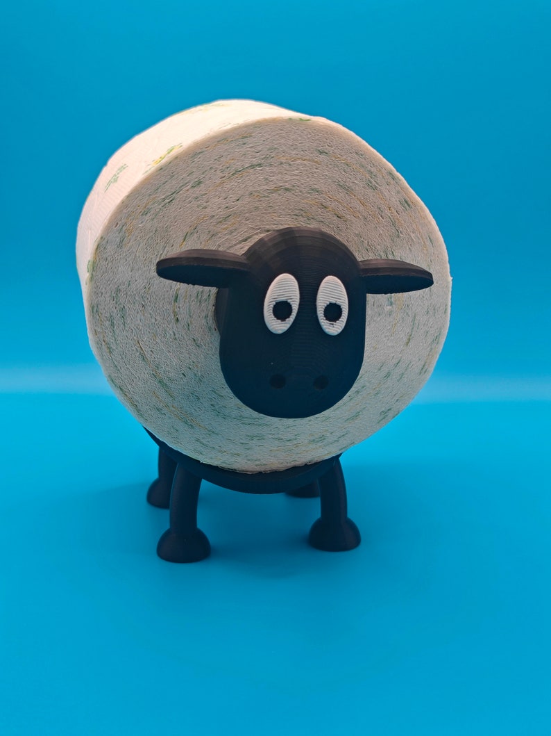 May include: A black sheep-shaped toilet paper holder with a white roll of toilet paper. The sheep has a black face with two white eyes.