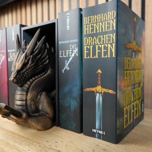 May include: A bronze-colored dragon bookend holds several books with titles in German. The dragon is sculpted with intricate details, appearing to read a book. The book spines are dark with gold lettering, featuring the titles "Die Elfen" and "Drachen Elfen".
