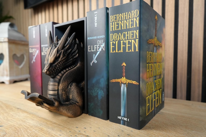 May include: A bronze-colored dragon bookend holds several books with titles in German. The dragon is sculpted with intricate details, appearing to read a book. The book spines are dark with gold lettering, featuring the titles "Die Elfen" and "Drachen Elfen".
