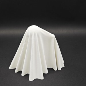 Puede incluir: Un objeto blanco de tela con forma de fantasma con una parte superior lisa y redondeada. El objeto est&aacute; drapeado sobre una superficie negra.