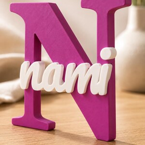 May include: A large, magenta-colored capital letter N made of a solid material. Attached to the front of the letter is the name nami written in white, lowercase, cursive-style letters. The object has a smooth, matte finish and stands upright.