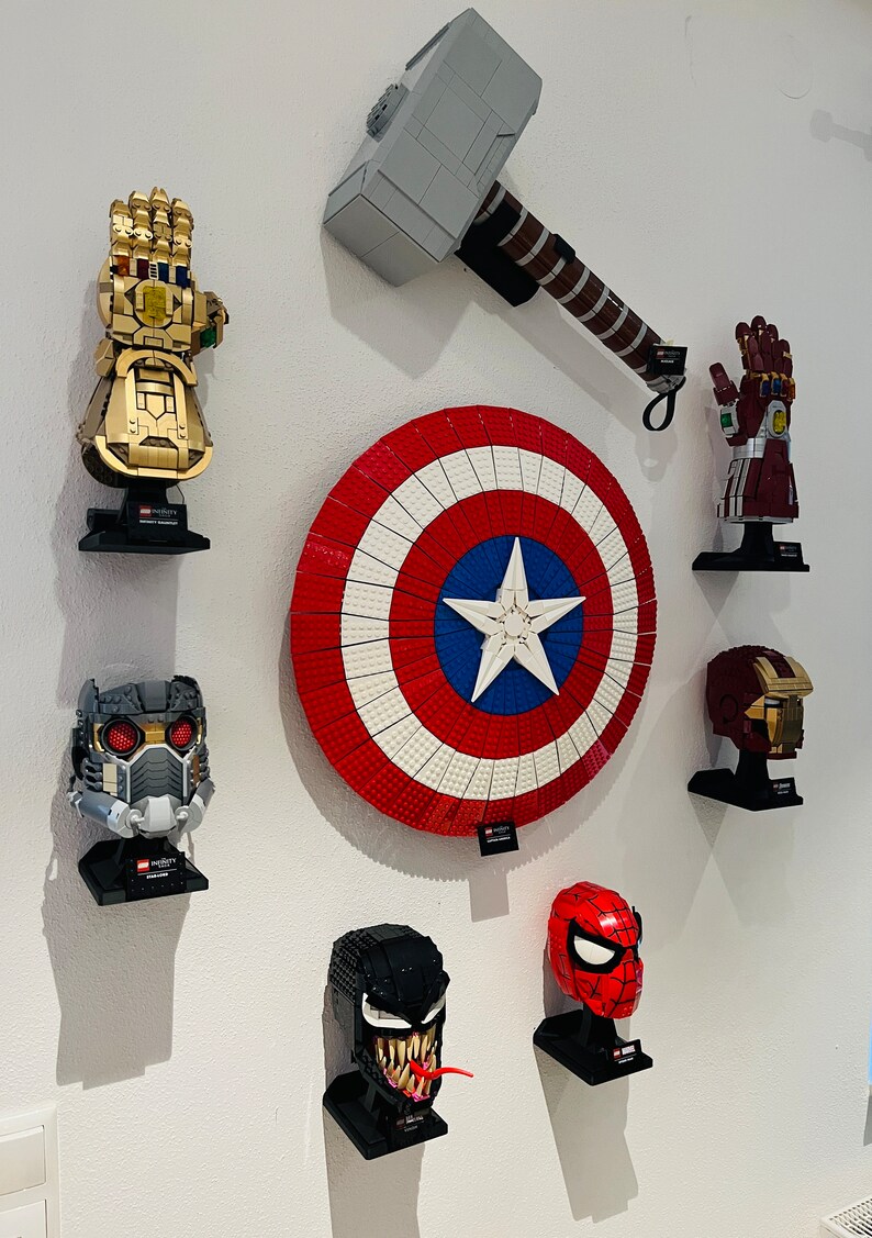 Op de afbeelding: Een verzameling Marvel-thema LEGO sets gemonteerd op een witte muur. Inclusief een gouden Infinity Gauntlet, Captain America's schild, Thor's hamer en verschillende karakterhelmen zoals Spider-Man en Venom.