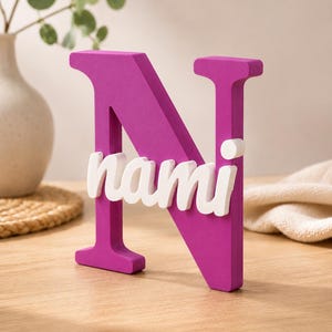 May include: A large, magenta-colored capital letter N made of a solid material. Attached to the front of the letter is the name nami written in white, lowercase, cursive-style lettering. The object stands upright on a flat surface.