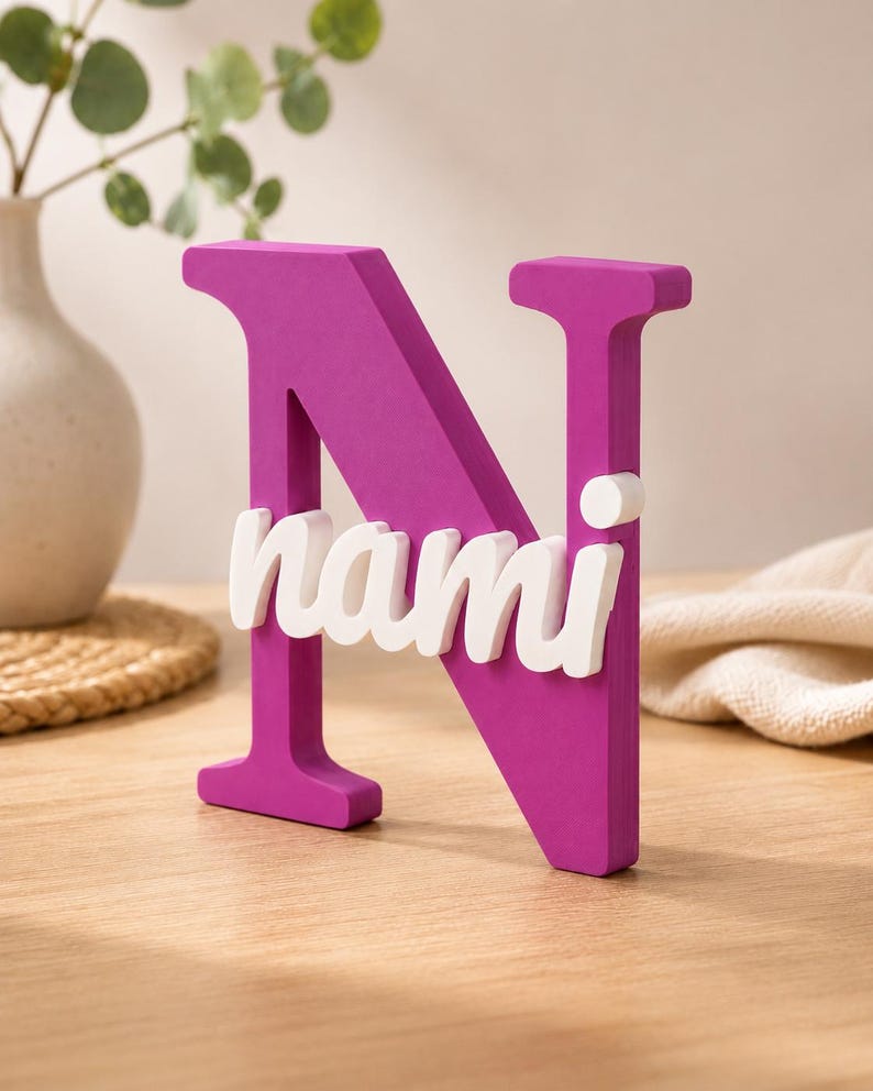 May include: A large, magenta-colored capital letter N made of a solid material. Attached to the front of the letter is the name nami written in white, lowercase, cursive-style lettering. The object stands upright on a flat surface.