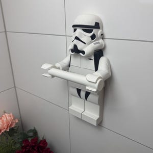 May include: White plastic toilet roll holder shaped like a Star Wars Stormtrooper. The holder is mounted on a white tiled wall.