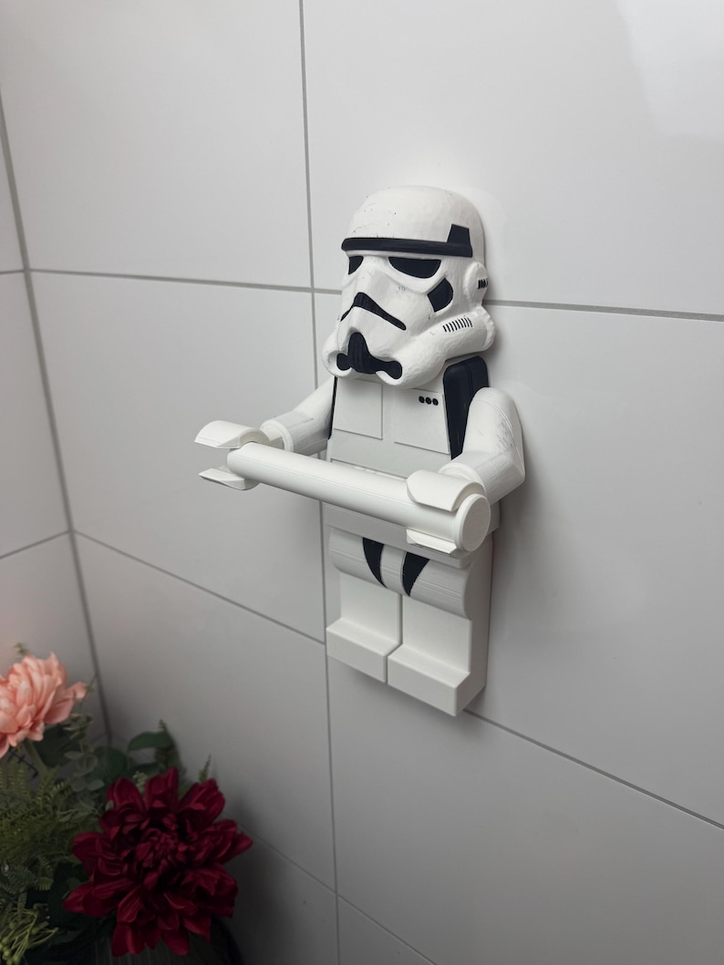 May include: White plastic toilet roll holder shaped like a Star Wars Stormtrooper. The holder is mounted on a white tiled wall.