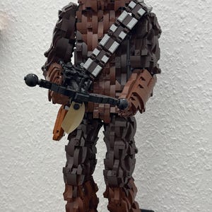 May include: A large Lego figure of Chewbacca from Star Wars, constructed from various shades of brown Lego bricks. The figure holds a black crossbow and stands on a black base with a small informational plaque. The figure is approximately 24 inches tall.