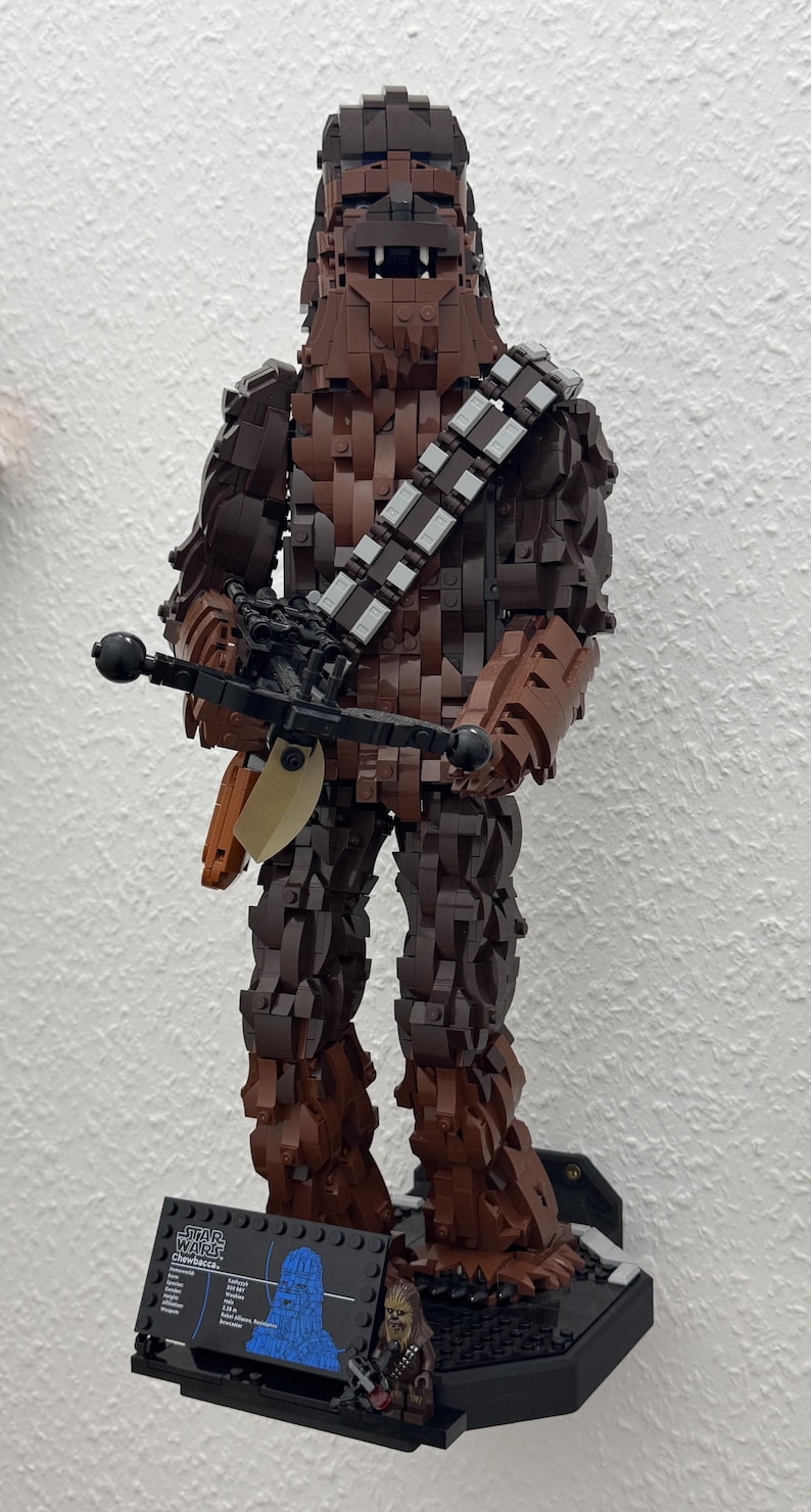 May include: A large Lego figure of Chewbacca from Star Wars, constructed from various shades of brown Lego bricks. The figure holds a black crossbow and stands on a black base with a small informational plaque. The figure is approximately 24 inches tall.