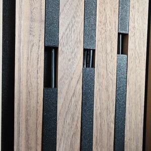 May include: Close-up of a wooden panel with vertical wooden slats and black textured strips. The panel has rectangular cutouts with black rods inside. The wood has a natural grain and colour.
