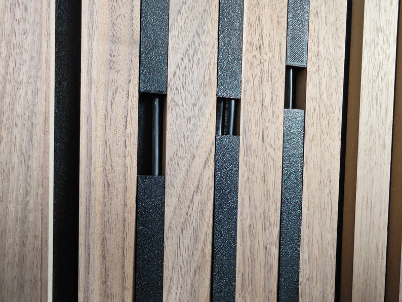 May include: Close-up of a wooden panel with vertical wooden slats and black textured strips. The panel has rectangular cutouts with black rods inside. The wood has a natural grain and colour.