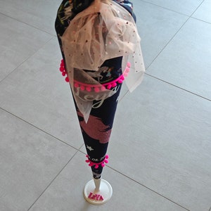 Puede incluir: Un cono escolar de unicornio negro y rosa con pompones rosas y un lazo de tul. El cono est&aacute; decorado con estrellas y la fecha "08. 2021". El cono est&aacute; sobre un soporte blanco con el nombre "Leya" escrito en letras rosas.