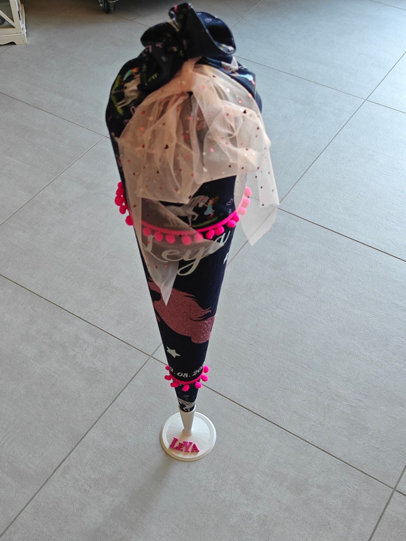 Puede incluir: Un cono escolar de unicornio negro y rosa con pompones rosas y un lazo de tul. El cono est&aacute; decorado con estrellas y la fecha "08. 2021". El cono est&aacute; sobre un soporte blanco con el nombre "Leya" escrito en letras rosas.