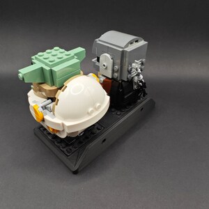Może przedstawiać: Lego minifigurka Mandaloriana stojąca obok Lego minifigurki Grogu w jego unoszącej się kapsule. Figurki znajdują się na czarnej podstawie Lego.
