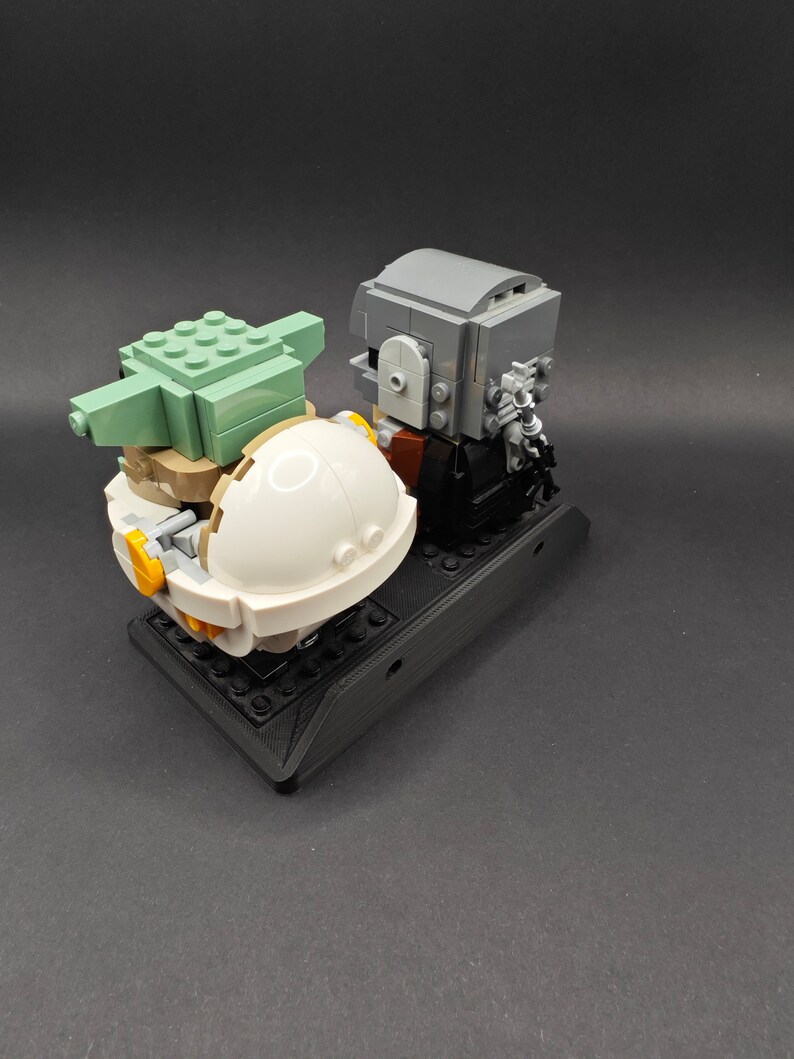 Może przedstawiać: Lego minifigurka Mandaloriana stojąca obok Lego minifigurki Grogu w jego unoszącej się kapsule. Figurki znajdują się na czarnej podstawie Lego.