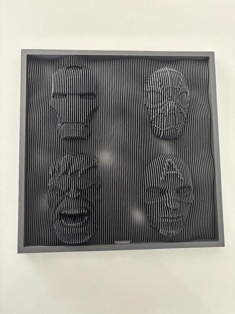 May include: A black framed wall art piece with three faces sculpted in a 3D style. The faces are made of thin, vertical lines that create a textured effect.