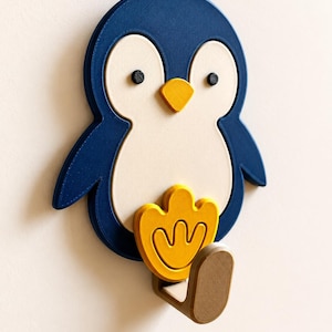 May include: A wall-mounted hook shaped like a penguin with a dark blue body, a cream-colored belly, two small black eyes, and a yellow beak. A yellow penguin foot is positioned above a brown hook that extends outward from the bottom of the figure.