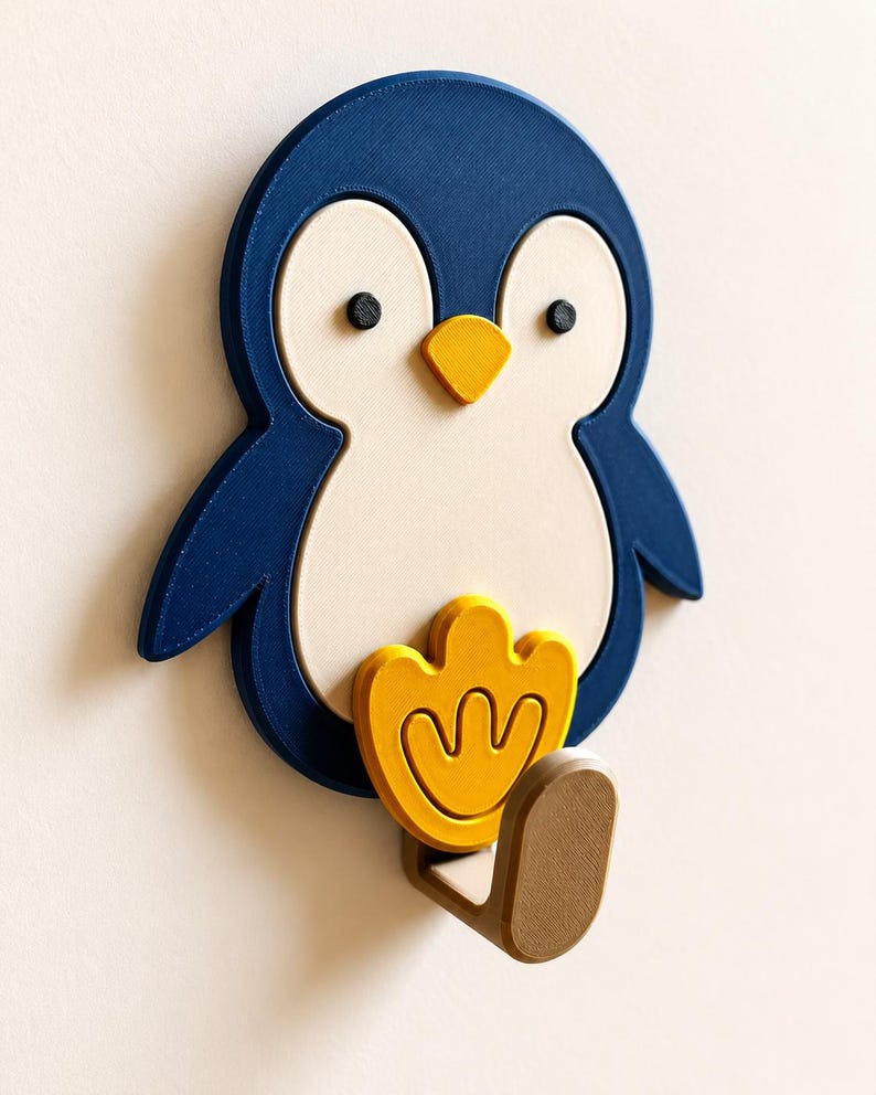 May include: A wall-mounted hook shaped like a penguin with a dark blue body, a cream-colored belly, two small black eyes, and a yellow beak. A yellow penguin foot is positioned above a brown hook that extends outward from the bottom of the figure.