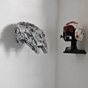 May include: A grey and white model of the Millennium Falcon spaceship is mounted on a white wall. Beside it is a white, red, and black pilot helmet on a black stand. The items are from the Star Wars franchise.