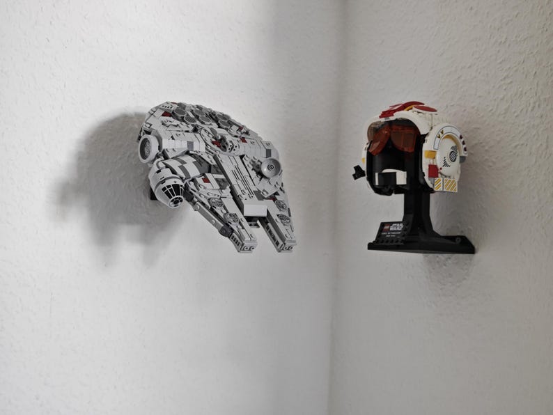 May include: A grey and white model of the Millennium Falcon spaceship is mounted on a white wall. Beside it is a white, red, and black pilot helmet on a black stand. The items are from the Star Wars franchise.
