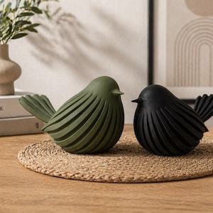 May include: Two decorative bird sculptures, one olive green and one black, sit on a woven mat. The birds have a stylized design with carved lines. The background includes a vase with greenery and a framed artwork.