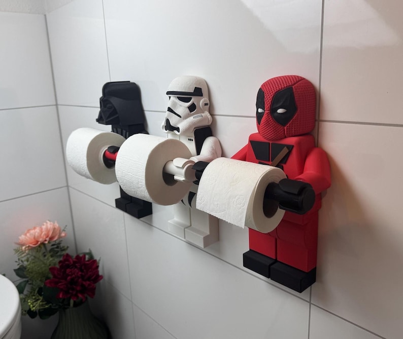 May include: Three toilet paper holders shaped like popular characters. A black Darth Vader, a white Stormtrooper, and a red Deadpool. Each holder has a roll of white toilet paper.