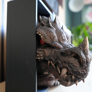 May include: A detailed, brown dragon head sculpture with sharp teeth and horns emerges from a black rectangular frame. The dragon has red eyes and a textured, scaly appearance, creating a fantasy theme.