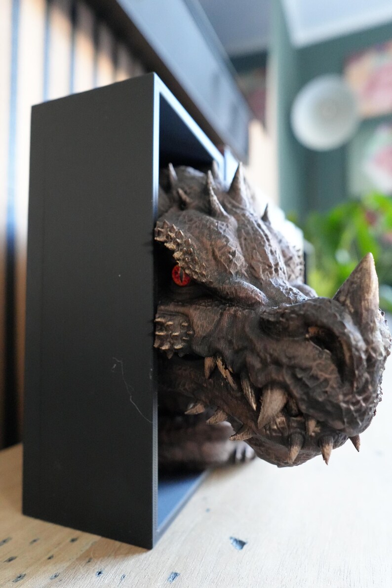 May include: A detailed, brown dragon head sculpture with sharp teeth and horns emerges from a black rectangular frame. The dragon has red eyes and a textured, scaly appearance, creating a fantasy theme.