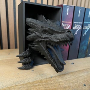 May include: A black dragon bookshelf decor, with the dragon's head and claws emerging from a black rectangular shelf. The dragon has detailed scales and teeth. Books with German titles are visible in the background.