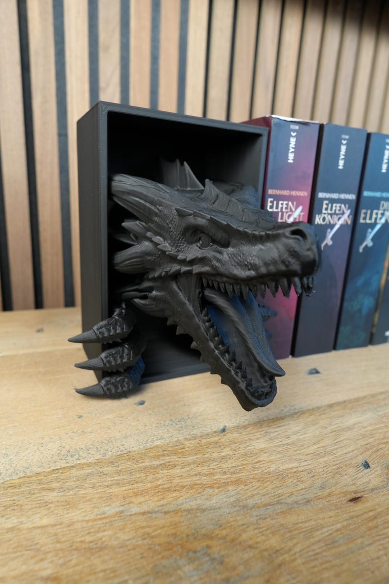 May include: A black dragon bookshelf decor, with the dragon's head and claws emerging from a black rectangular shelf. The dragon has detailed scales and teeth. Books with German titles are visible in the background.