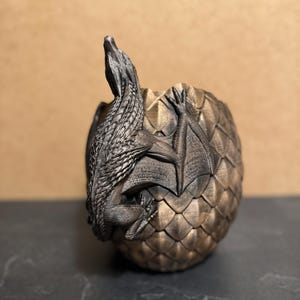 May include: A bronze-colored dragon figurine emerging from a textured egg. The dragon has detailed scales and wings, with its head and neck extended upwards. The egg has a geometric pattern.