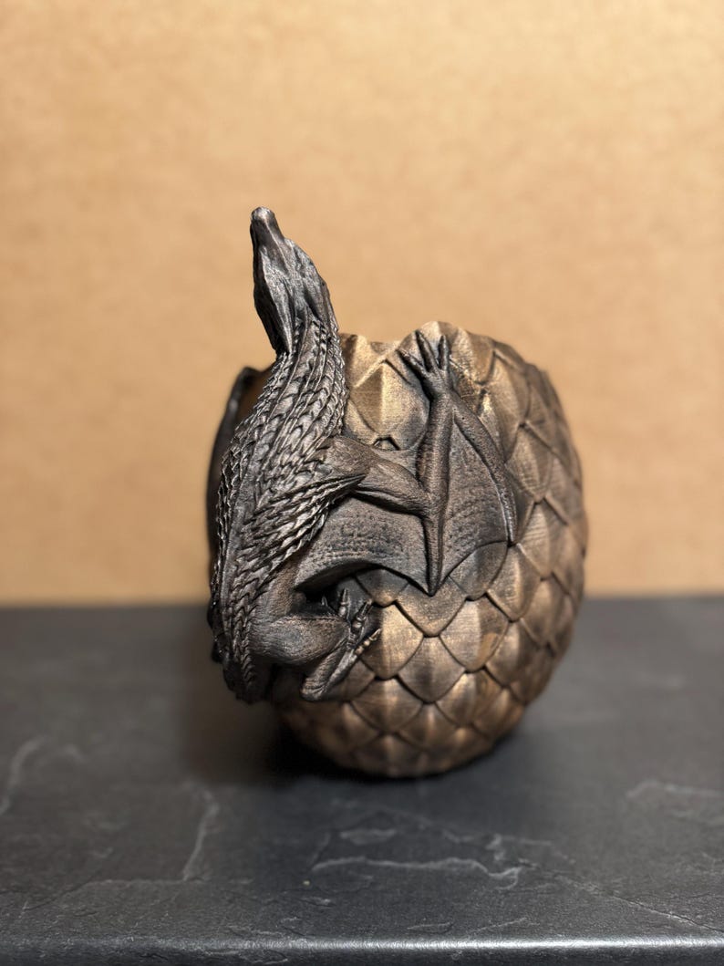 May include: A bronze-colored dragon figurine emerging from a textured egg. The dragon has detailed scales and wings, with its head and neck extended upwards. The egg has a geometric pattern.