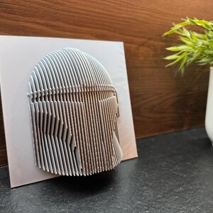 Puede incluir: Una escultura de pared de arte 3D impresa en blanco de un casco. El casco est&aacute; hecho de muchas l&iacute;neas verticales delgadas que crean un efecto texturizado.