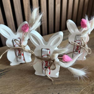 May include: Three white bunny-shaped figurines with a chocolate bar inside each. The bunnies are decorated with pink and white dried flowers and brown twine.